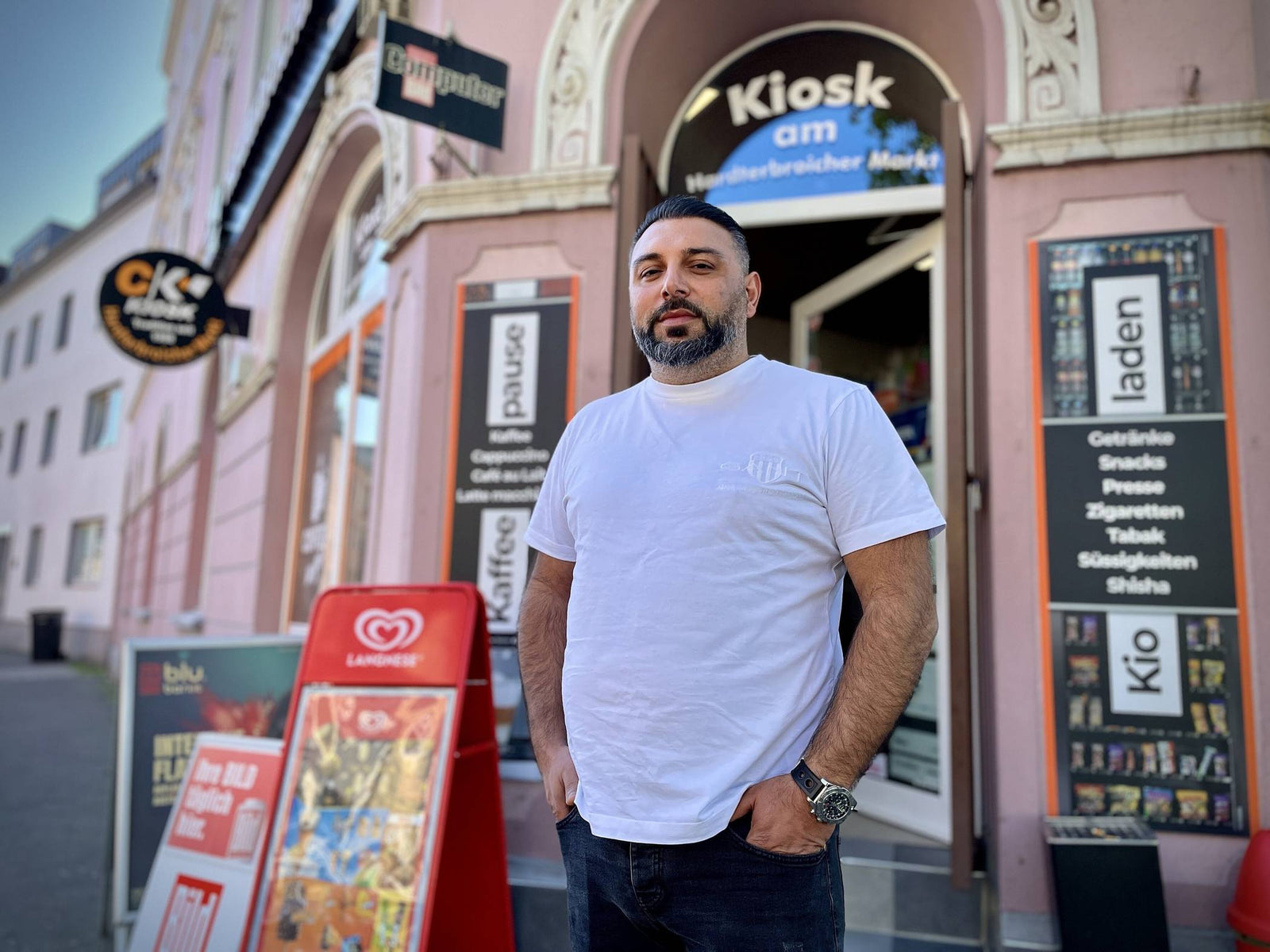 Kioskbetreiber in weißm T-Shirt steht vor seinem Laden